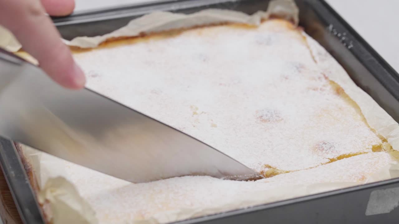 Nærbilde av en skarp kniv som skjærer gjennom fersk sitronkake med melisdryss i en bakeform