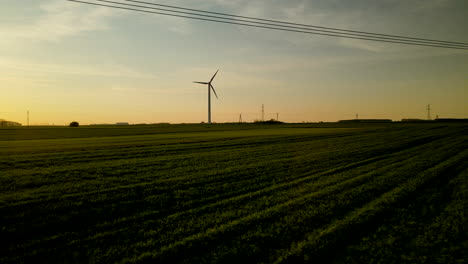 Drones-De-ángulo-Bajo-Vuelan-Sobre-Campos-Verdes-Al-Atardecer,-Silueta-De-Turbina-Eólica-En-El-Fondo