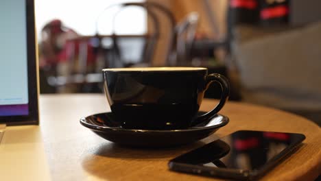 moviéndose hacia atrás desde una taza de té negra en un platillo y un teléfono inteligente a su lado en la mesa redonda de madera, disparo dinámico de dolly