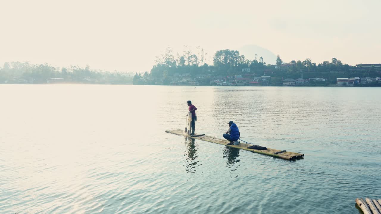 Indonesiske fiskere klar til at kaste et bredt net fra en simpel bambusflåde på en rolig sø med spejling