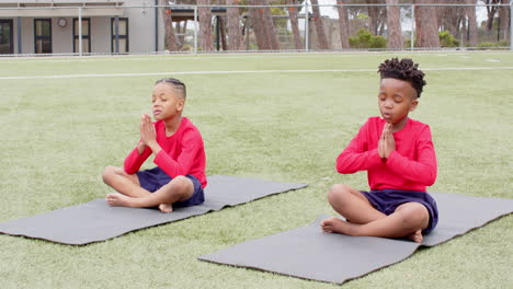 multiracial boys practicing yoga on mats on school field, meditating with eyes closed