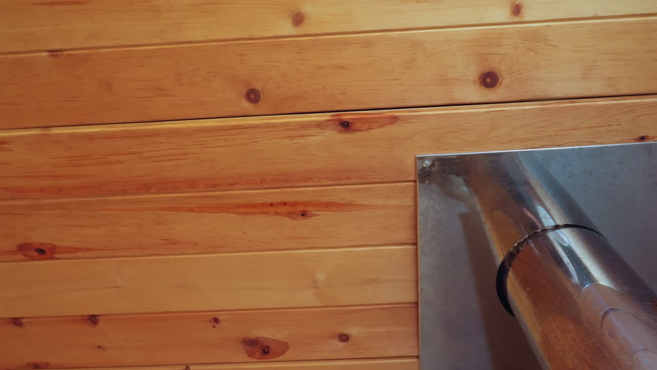 Close up of wooden ceiling with shiny metal stove pipe passing through fitted square panel, displaying warm wooden textures, grain, knots, and structural detail in cozy rustic interior setting