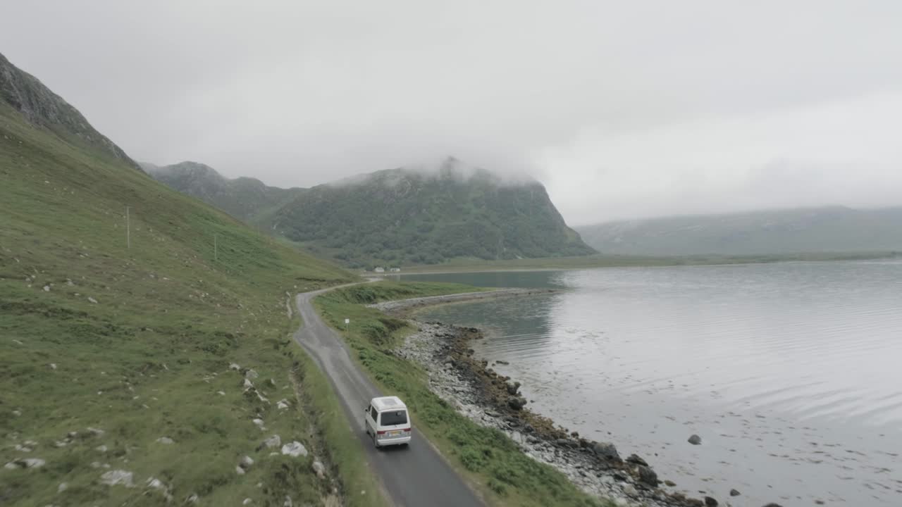 Close up drone tracking shot of a campervan on a single track road during a trip of the Scotland North Coast 500 with a Loch and cloudy topped mountains in view to passing the camper van