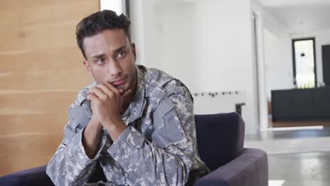 thoughtful biracial male soldier in uniform sitting in armchair at home, slow motion