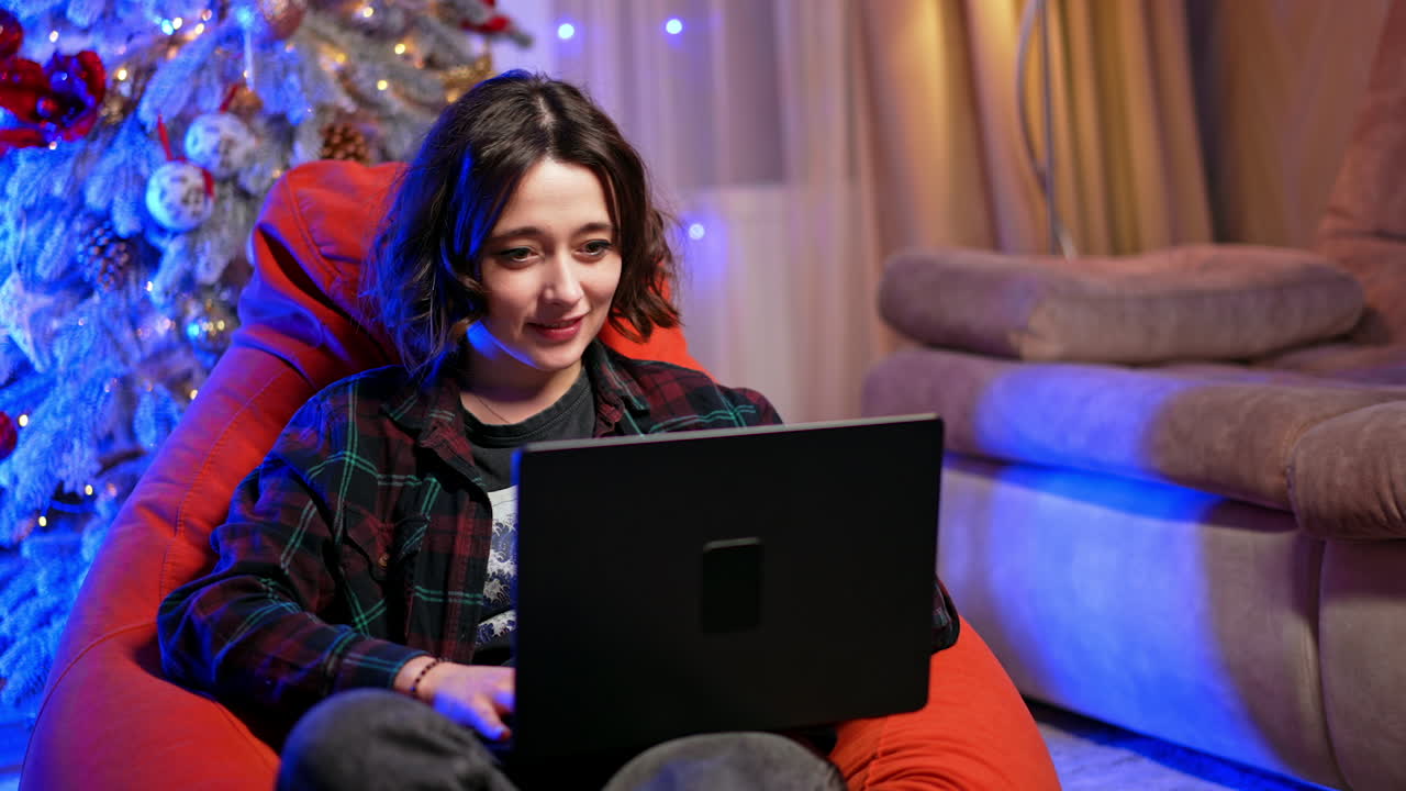Smiling girl sitting in the bean bag chair with laptop. Work from home remote online. Christmas tree at backdrop.