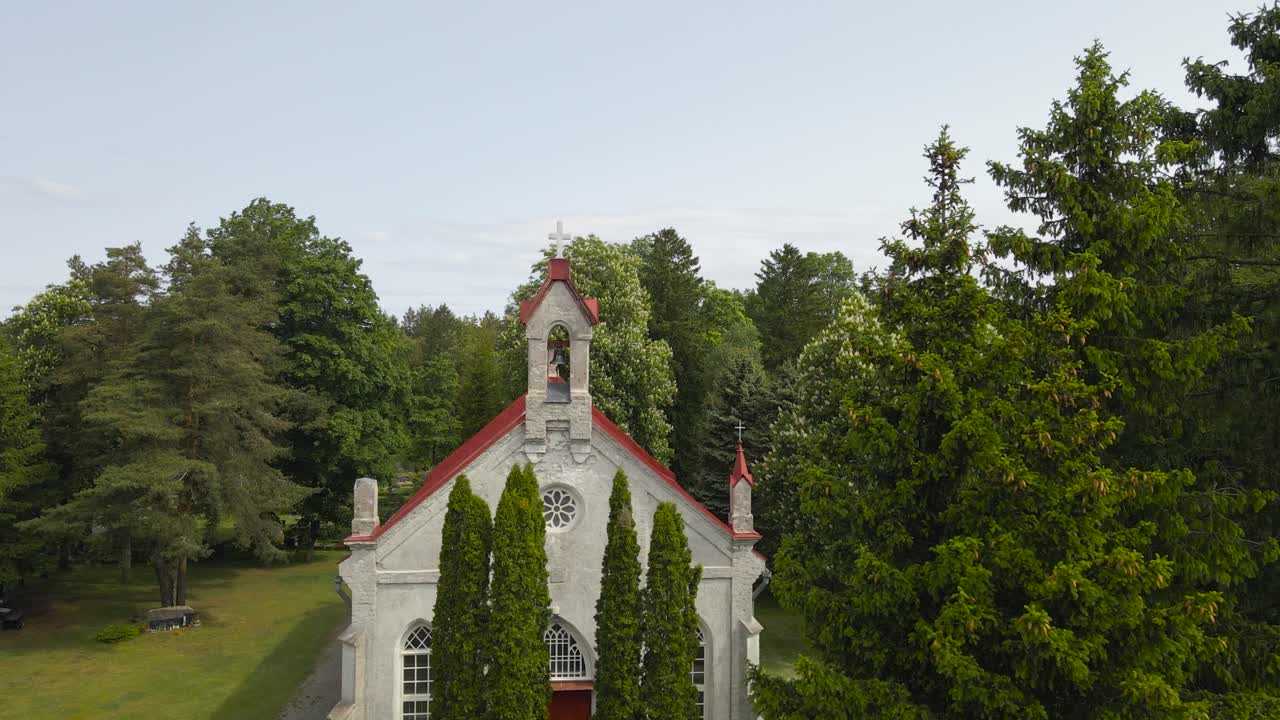 Gorgeous aerial drone footage orbiting around a large old historic white colored church with red roof and crosses that has a church bell in the countryside during a warm summer day. Trees in front.