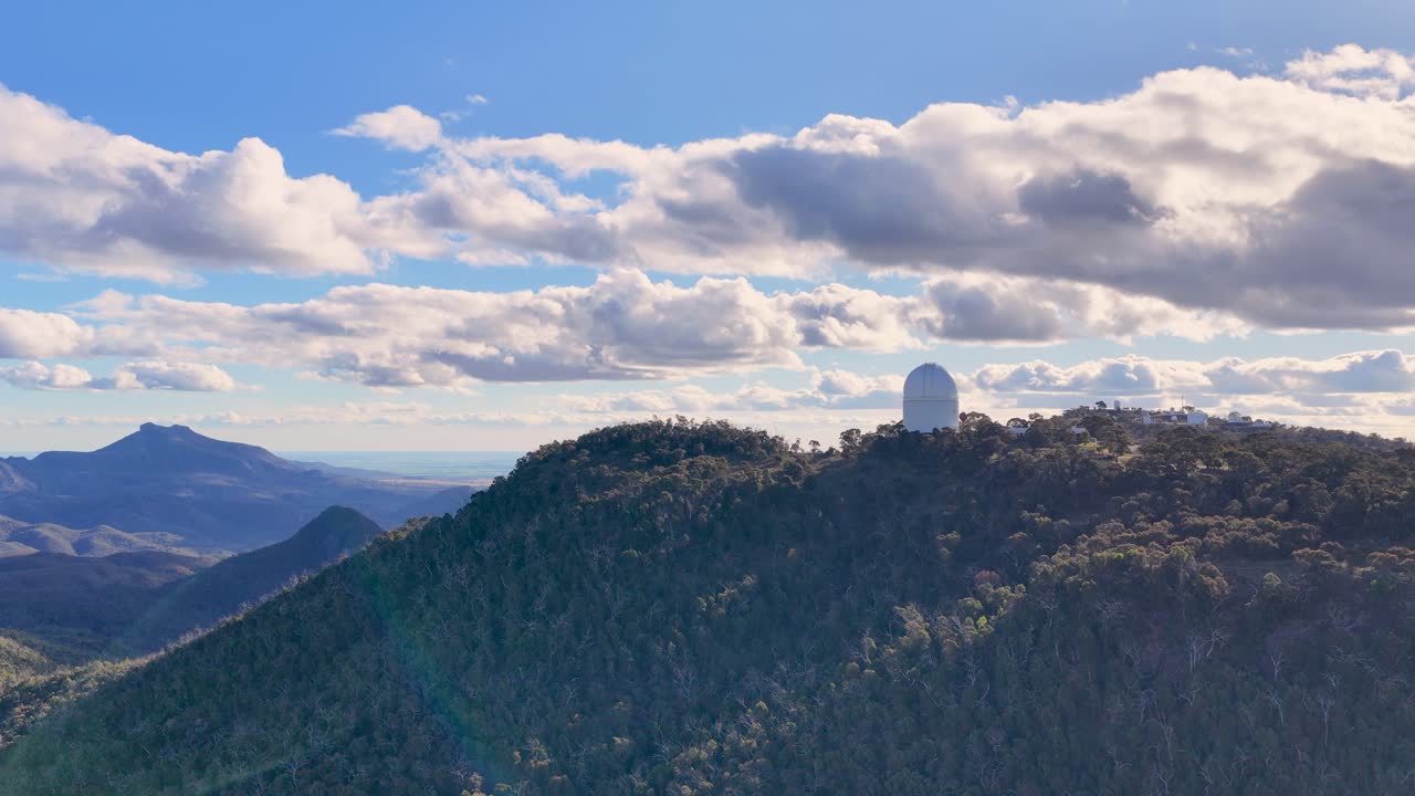 Drone footage pans across a forested mountain ridge with a large observatory dome under partly cloudy skies, capturing scenic Australian landscape in daylight