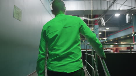 back shot of male walking on stairs, holding rail while heading towards billiard room. green lighting casts dramatic effect, modern interior design adds depth