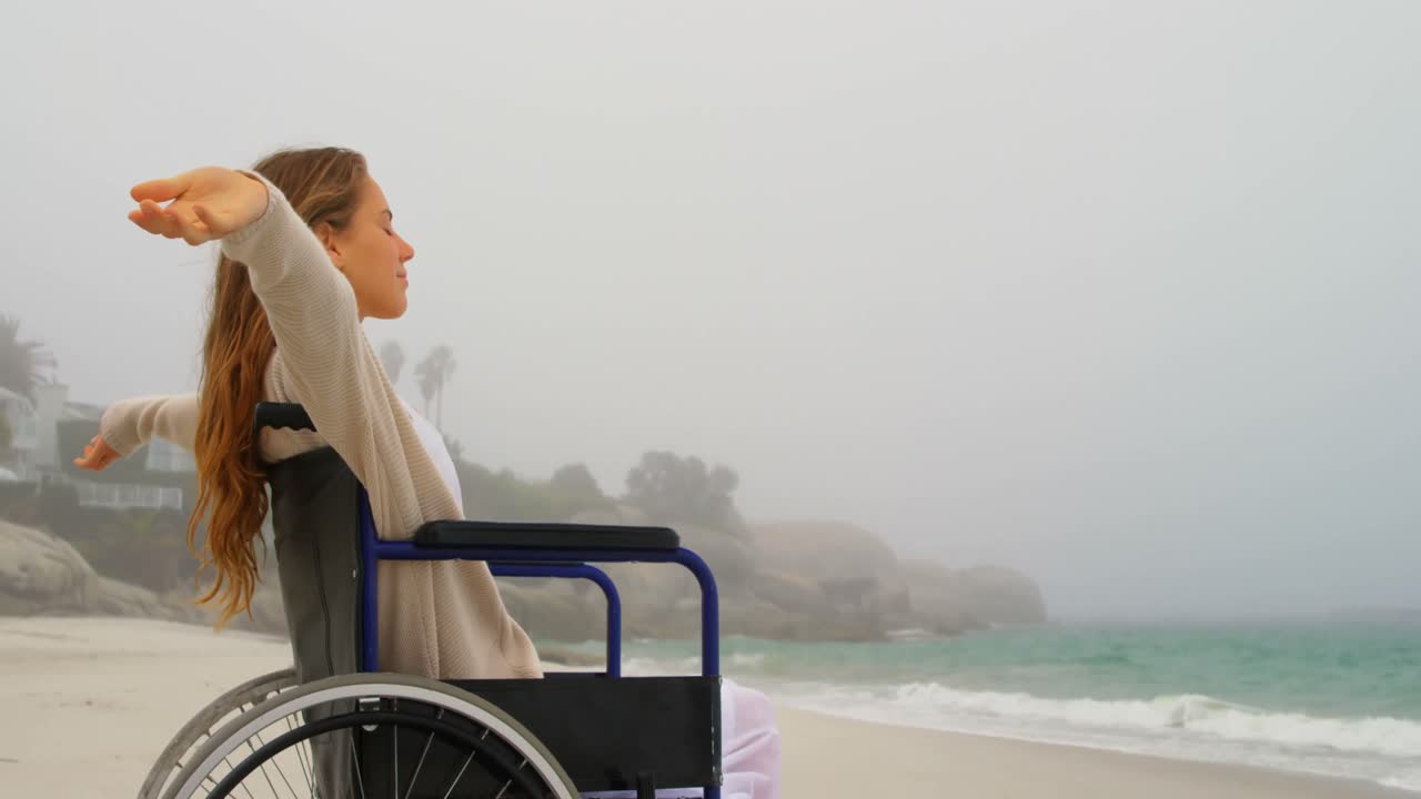 Side view of young Caucasian woman sitting with arms outstretched on wheelchair at beach 4k