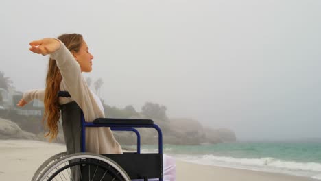 side view of young caucasian woman sitting with arms outstretched on wheelchair at beach 4k