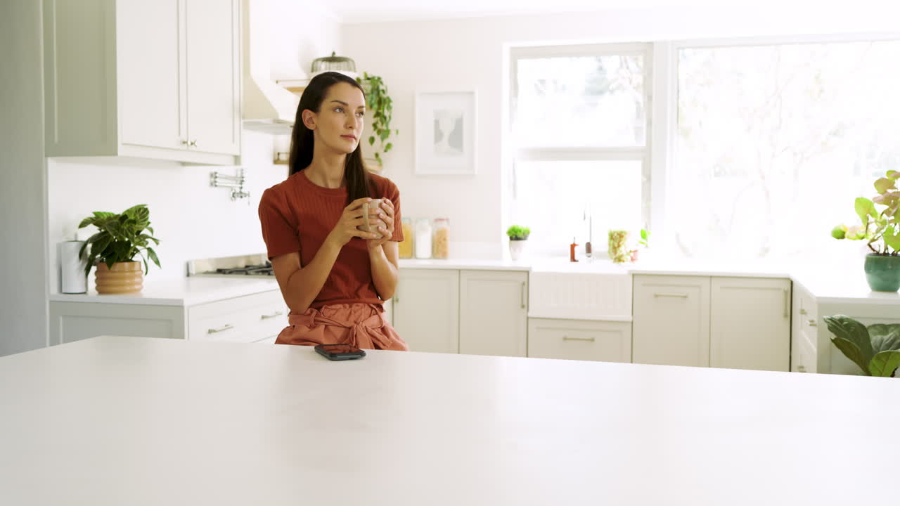Donna in cucina che usa lo smartphone, sorseggia un caffè e si rilassa a casa.