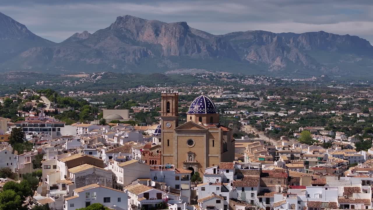 Drone circles around Nuestra Señora de Consuelo church with iconic blue dome, white houses, hills, and trees in Altea, Spain.