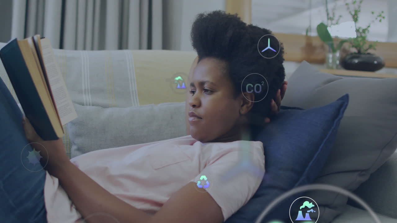 Animation of eco icons and data processing over african american woman reading book on sofa at home