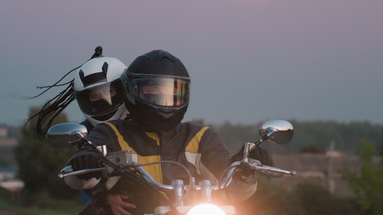 Close up of motorcyclist in black helmet and leather jacket riding uphill with sibling passenger holding tight, both wearing safety helmets, reflections of sunset visible in visors