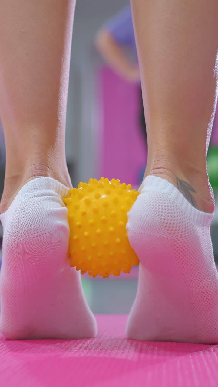 primer plano de una persona con una pierna tatuada, cubierta ligeramente por calcetines, sosteniendo una pelota amarilla puntiaguda bajo el pie para el ejercicio de acumulación de resistencia en una alfombra rosa
