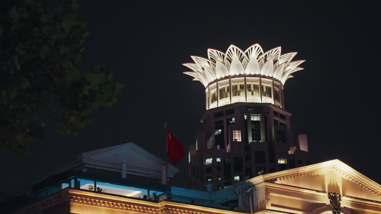 Gece çatının üzerinde aydınlatılmış taç kulesi, süslü taç yapısı karanlık gökyüzüne karşı parlıyor, ağaç silüeti ve kırmızı bayrak kontrast katıyor, sıcak korniş aydınlatması tarihi cepheyi vurguluyor, sinematik