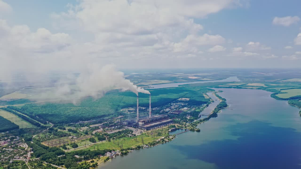 Huge manufactory plant among natural background. Smoke comes from pipes into the atmosphere near the river and green forest outdoors. Aerial view.