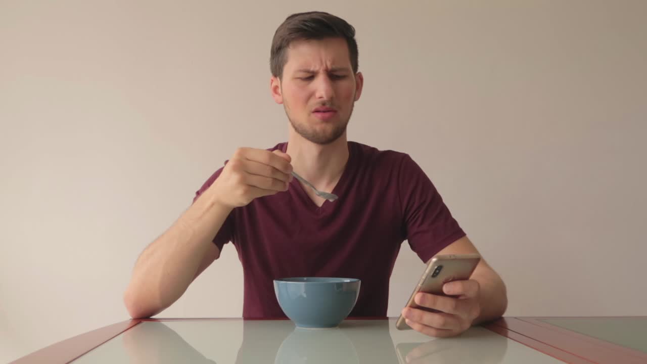 joven comiendo cereales y reaccionando al contenido del teléfono con disgusto y sorpresa, todavía en plano medio a la altura de los ojos frente a la cámara