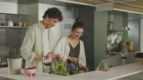 a happy couple cooking salad together in a modern kitchen with a laptop and wine
