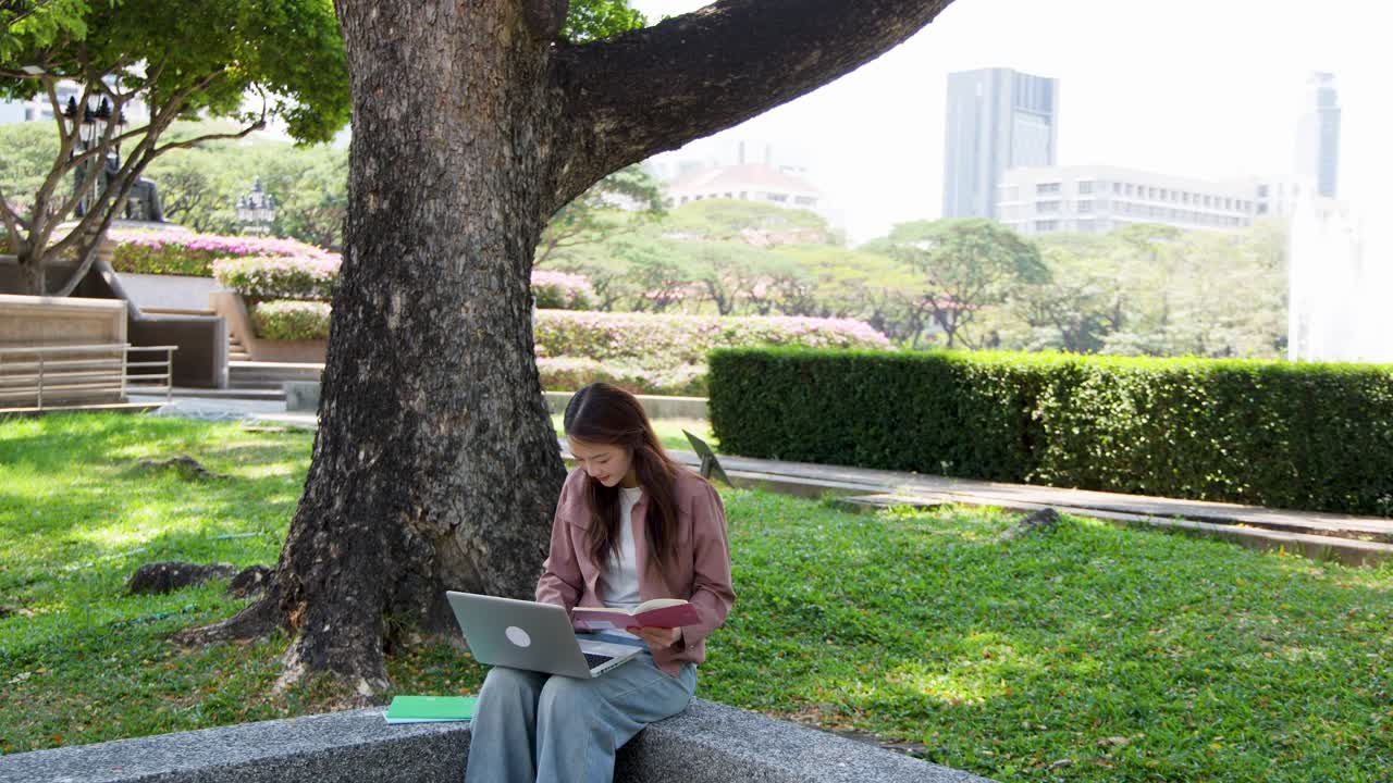 A young woman sits under a large tree using a laptop and reading a book