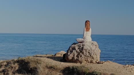 aerial view. flying the drone in a circle. a young woman in white clothes sits on a stone at the top of a hill against the background of the sea horizon. attainment. vertex.