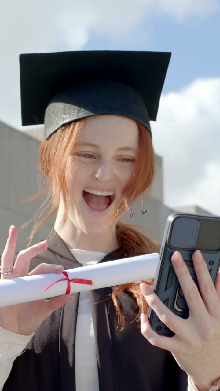Video verticale: Laureato sorridente e attento che tiene in mano diploma e telefono nell'atrio del campus, con tocco e toga.