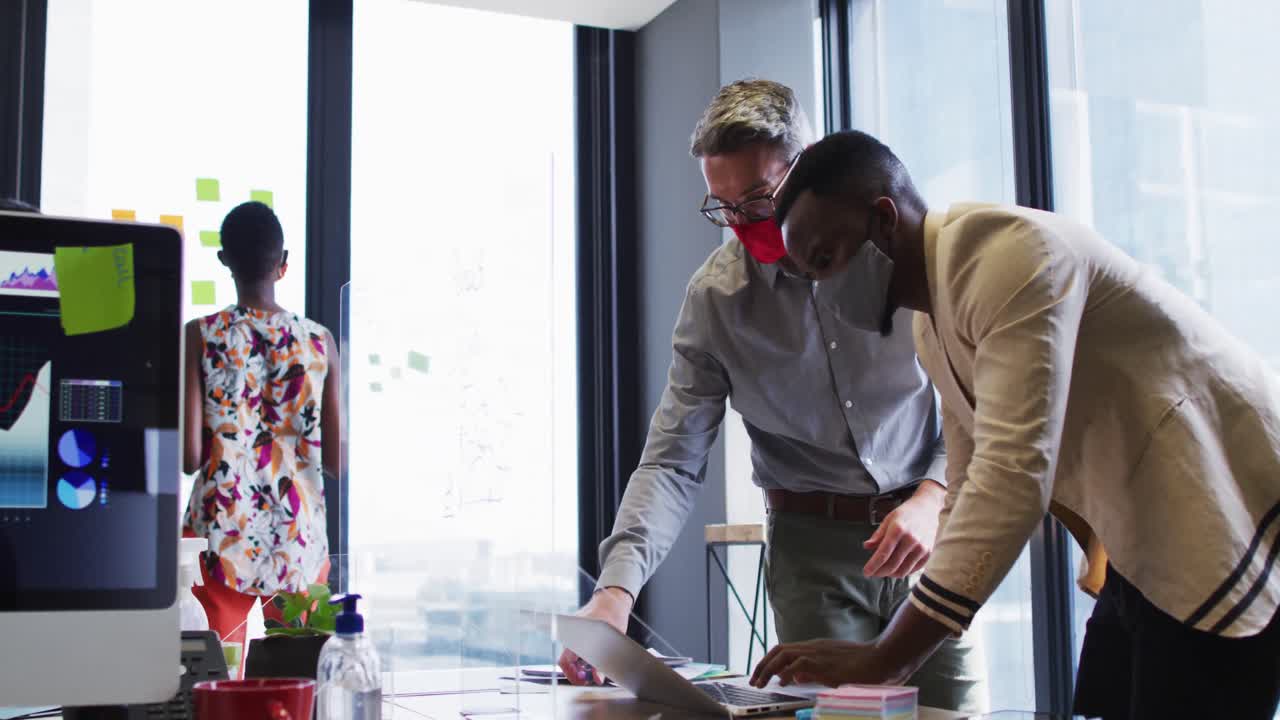 African american man and man wearing face masks discussing over laptop at modern office