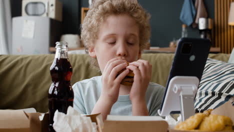 young boy eating a hamburger while watching a smartphone
