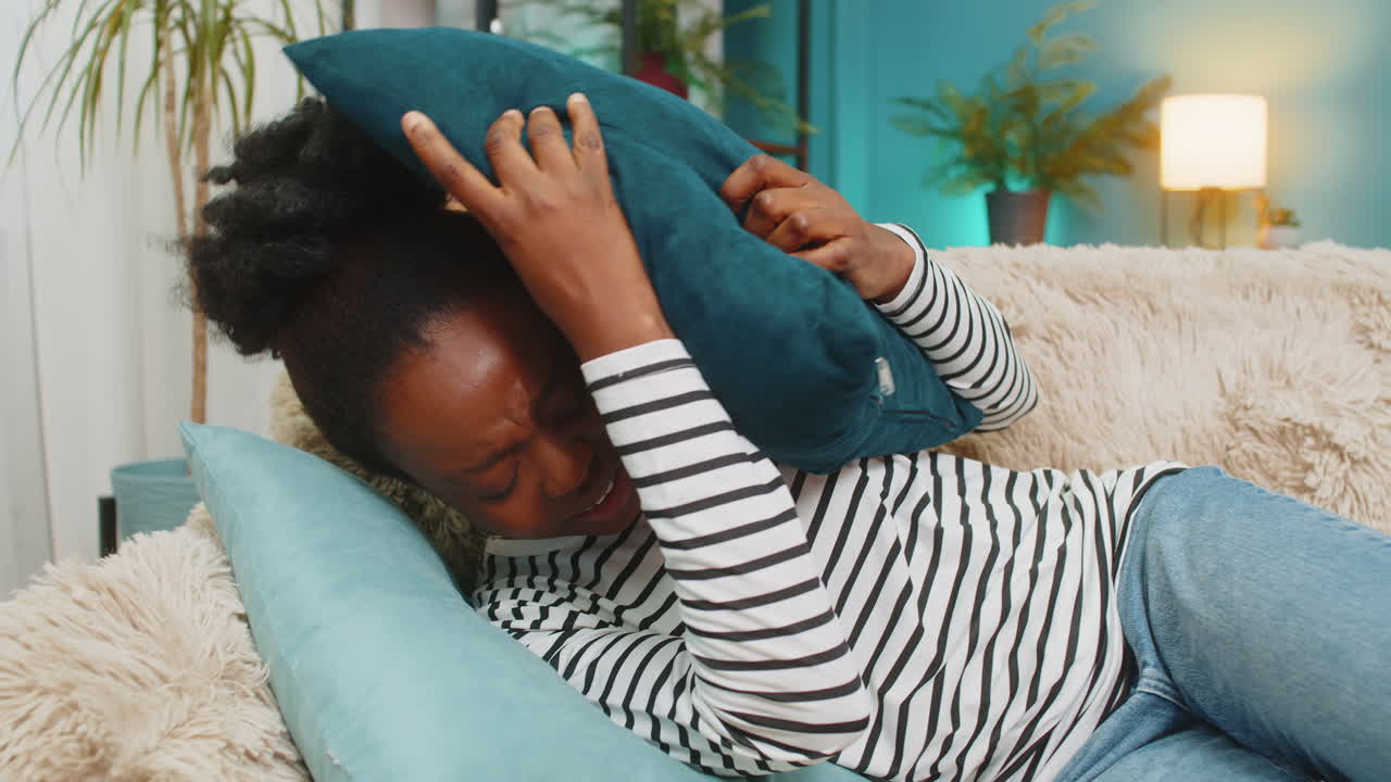 Afroamerikansk kvinde ligger på sofaen, lukker øjnene og dækker ansigtet med en pude, mens hun har migræne.