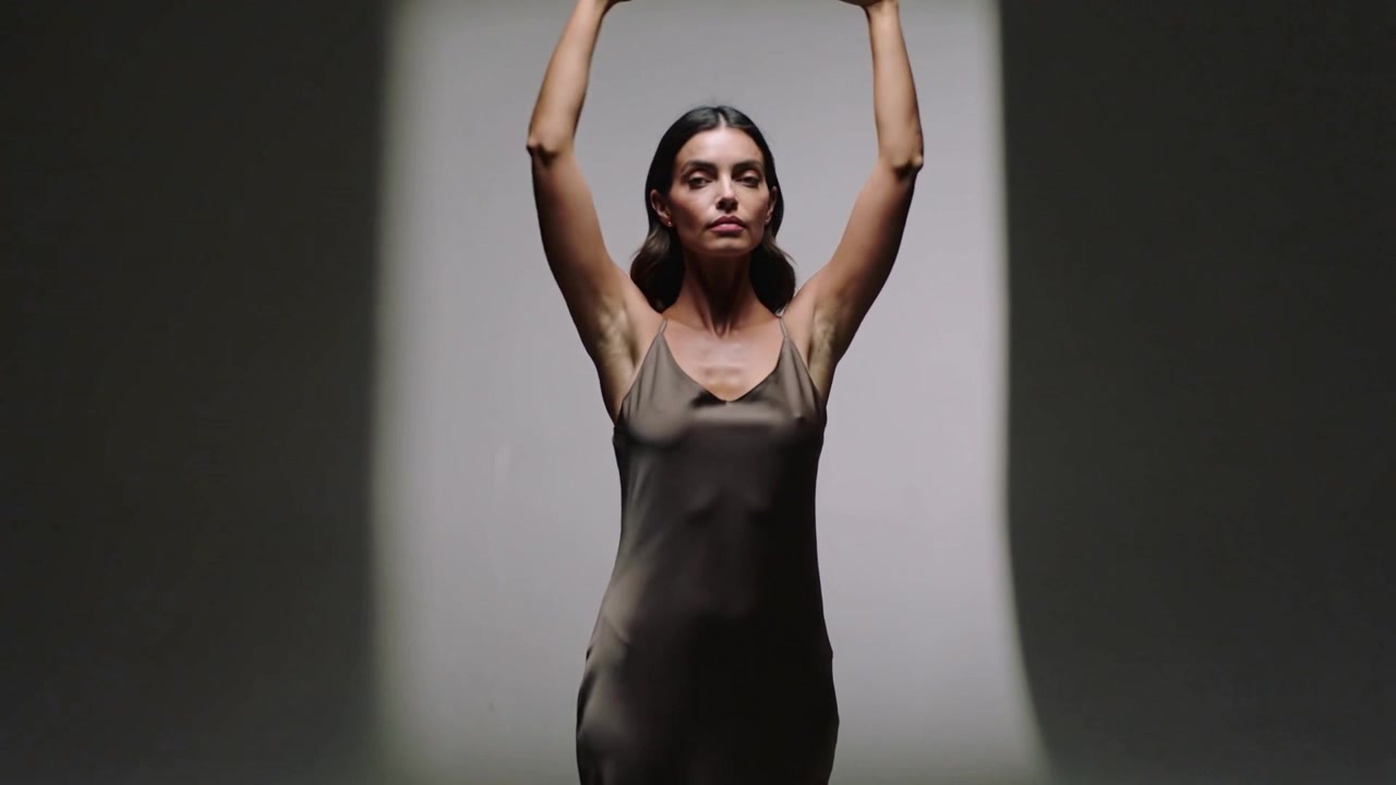 Shifting camera filming model raising arms and posing in studio with lightbox, satin dress