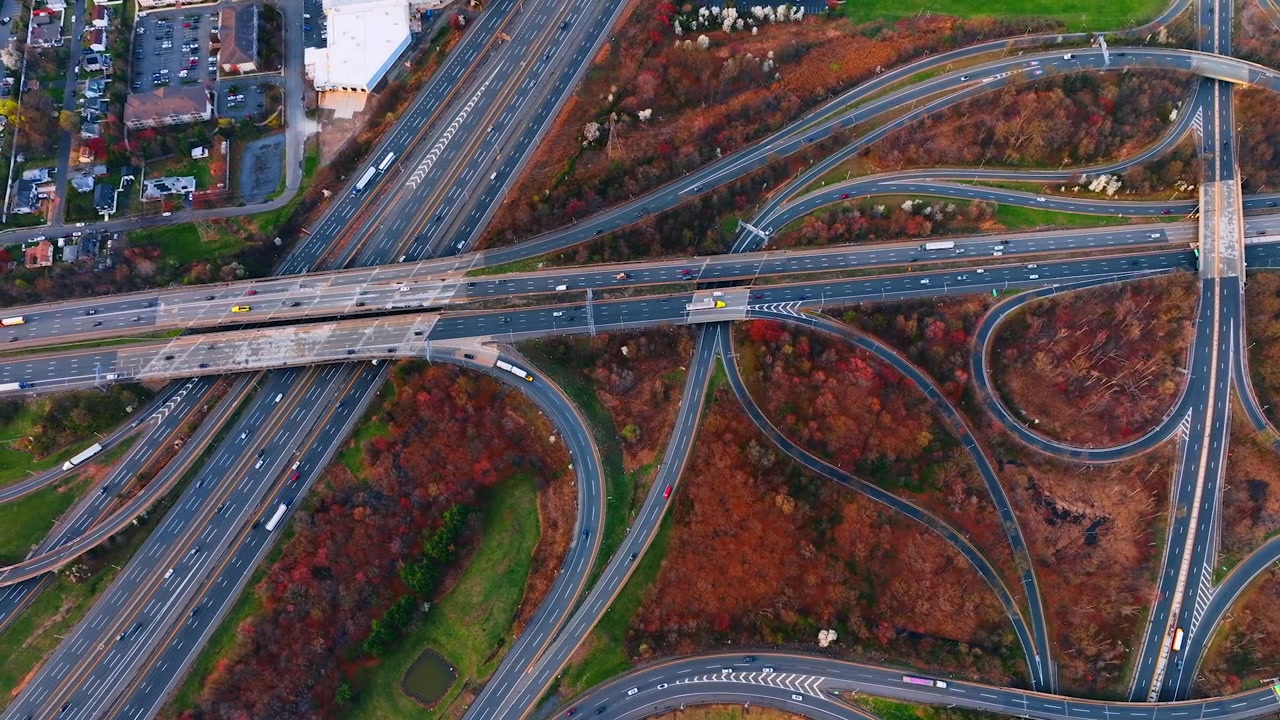 Top-down view of a highway interchange with autumn foliage, New Jersey. Overhead of curving ramps and an overpass crossing multilane roads between reddish woodland and a parking lot