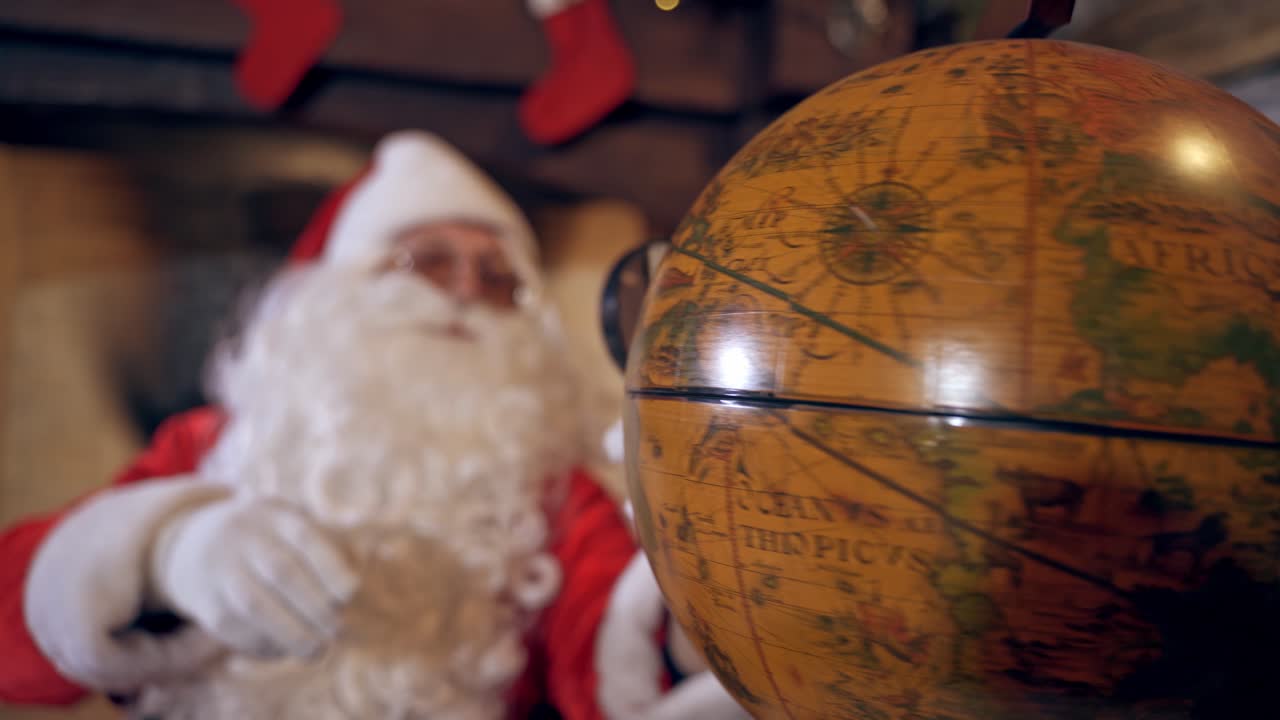 Model of globe on the table. Santa with white beard looking through the magnifying glass on the globe. Christmas time.