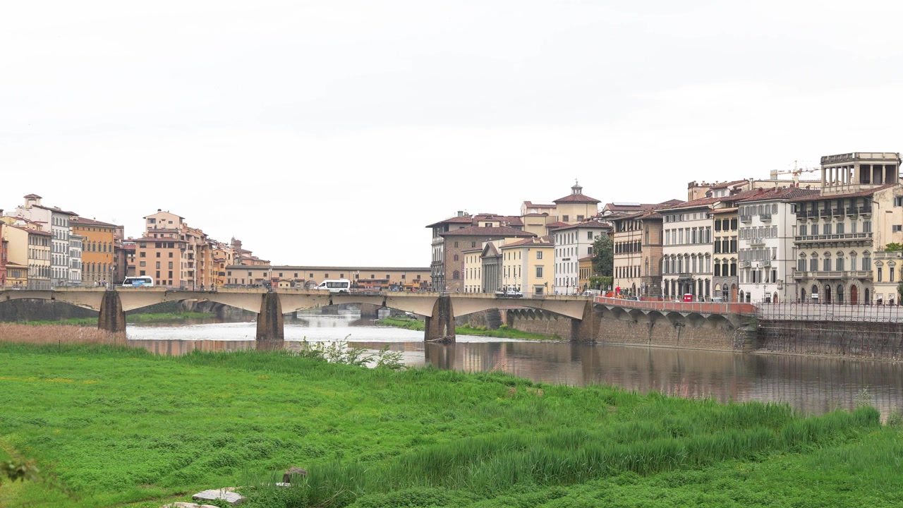 Ampia inquadratura stabile dell'iconico Ponte Vecchio di Firenze che attraversa il fiume Arno, incorniciato da rigogliose sponde verdi, edifici rinascimentali e luce naturale nuvolosa. Nessuna persona visibile.