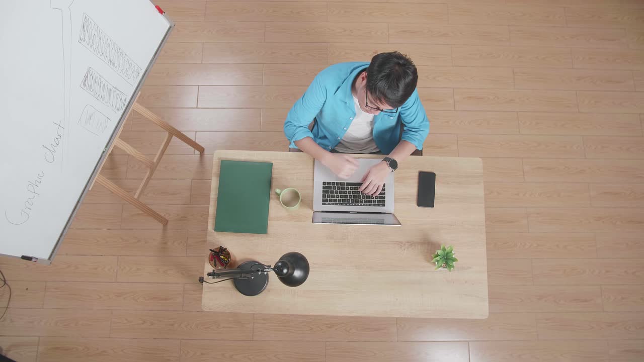 위쪽 뷰  아웃: 집에 있는 스마트폰 에 있는 노트북에 타입하는 화이트보드 에 앉아 있는 나무 책상에 앉은 채 지표손가락을 올리는 아시아 남자의 사진