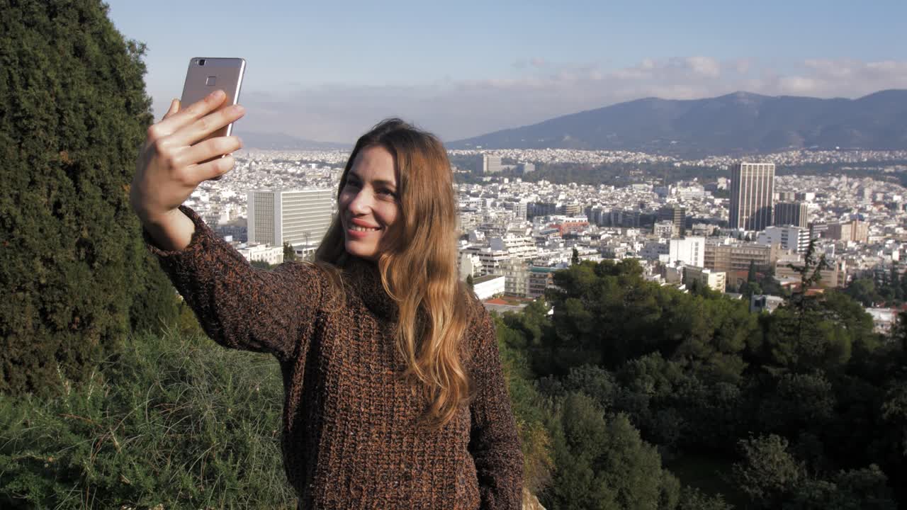 Young woman lifts her phone up to take a smiling selfie having city of Athens in the background