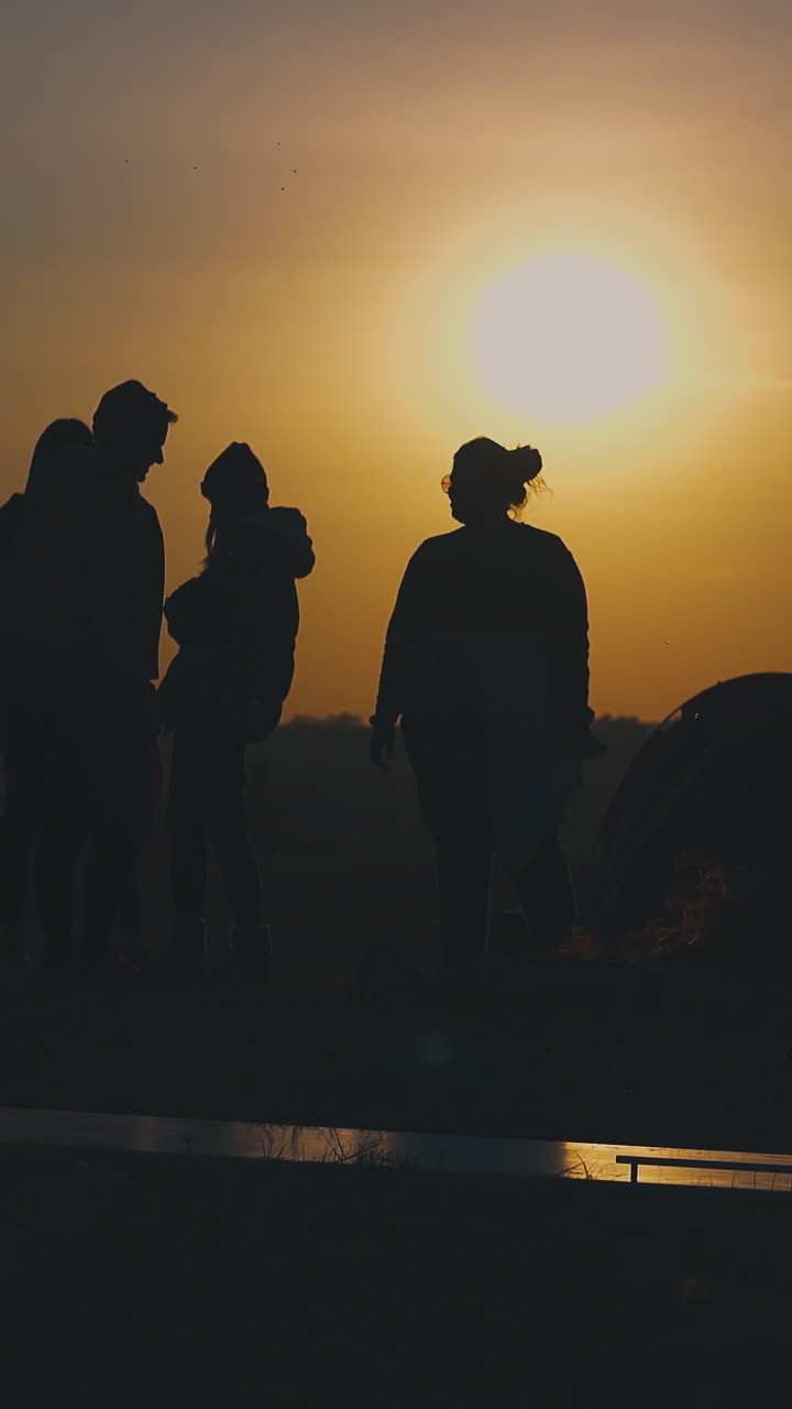 young people tourist silhouettes rest near burning bonfire and tent against clear evening sky with orange sunset