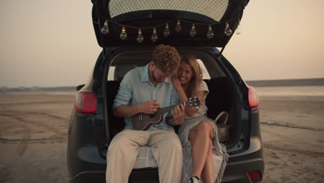 A-bearded-man-with-curly-hair-in-a-blue-shirt-is-playing-the-ukulele-and-his-blonde-girlfriend-is-hugging-him,-they-are-laughing-and-sitting-in-the-trunk-of-the-black-car-decorated-with-the-lights-against-the-yellow-sky,-while-their-dog-is-lying-on-the-sand