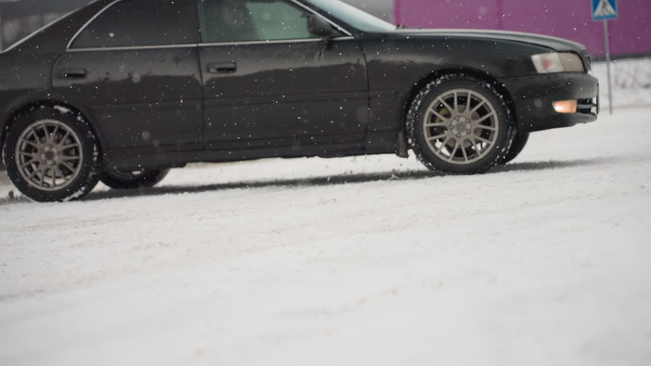 car driving through heavy snowfall on snow covered road kicks up slush and snowflakes spinning tires visible motion against blurred industrial fence and post under gray winter sky
