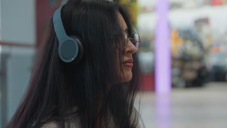 close up on young woman with long hair wearing headphones and glasses, sitting calmly with serene expression, focused downward, blurred indoor background
