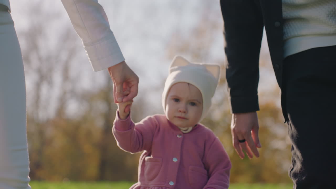 Tumling lærer at gå med forældre, et familiemoment i efterårspark, nærbillede af lille barn i lyserød jakke og strikhue, guidet af mors og fars hænder, varmt sollys på græsset, blid støtte.