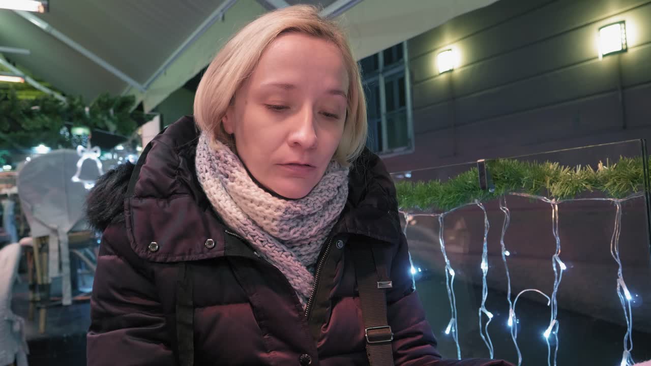 Woman eating in cafe, outdoors on terrace, turning away, talking - Christmas lights all around