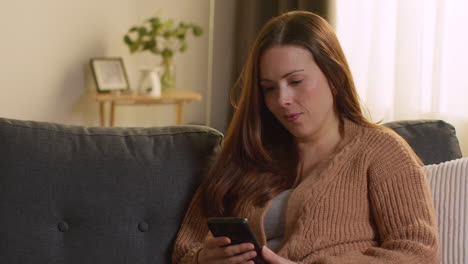 Smiling-Woman-Sitting-On-Sofa-At-Home-Using-Mobile-Phone-To-Check-Social-Media-Message-And-Scrolling-Online