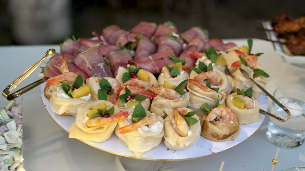 Platter features colorful appetizers including shrimp rolls, meat wraps, and bite-sized treats. Guests enjoy the tasty food in a relaxed outdoor setting