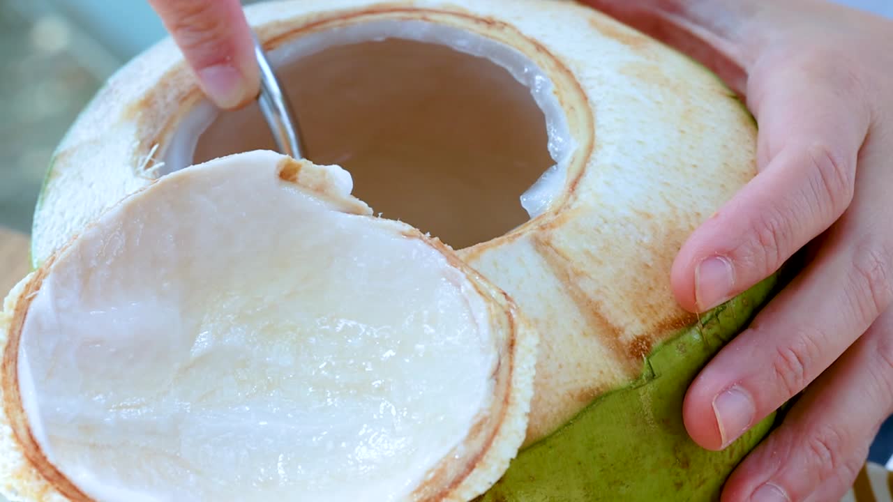 A hand uses a spoon to scoop fresh coconut meat from a green coconut shell.