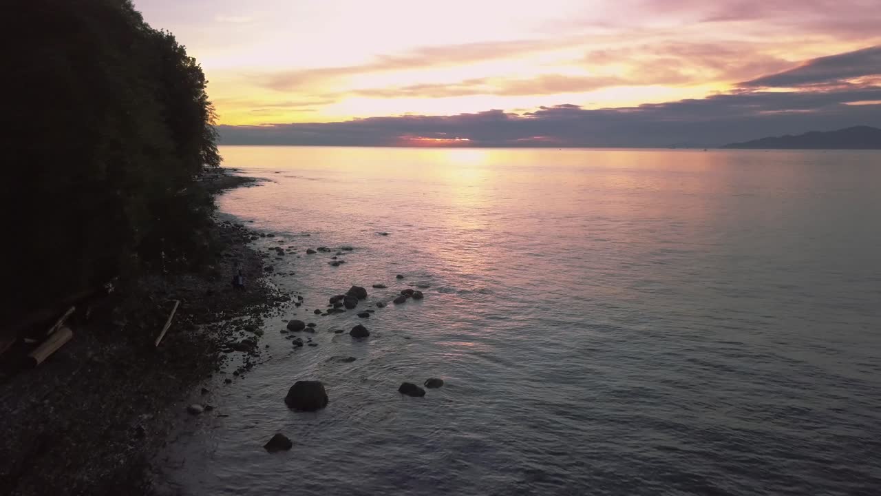 puesta de sol aérea tiro medio ancho volando sobre el océano pacífico y las rocas hacia el sol y la reflexión al lado de la playa rocosa con viejos árboles altos y madera a la deriva en vancouver, columbia británica, canadá