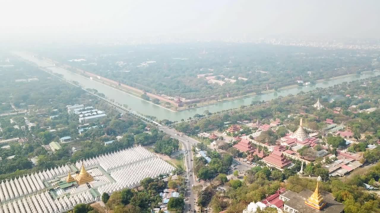 Aerial view captures the cityscape of Mandalay, Myanmar, showcasing a network of waterways, numerous buddhist temples and pagodas, and the dense urban sprawl extending towards the horizon