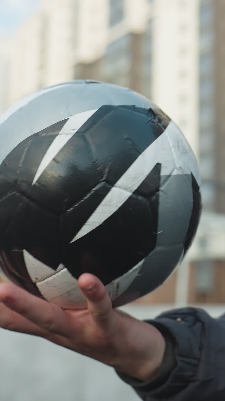 hombre habilmente gira la pelota de fútbol en su palma, mostrando equilibrio y enfoque, con edificios urbanos de gran altura y valla de malla en el fondo