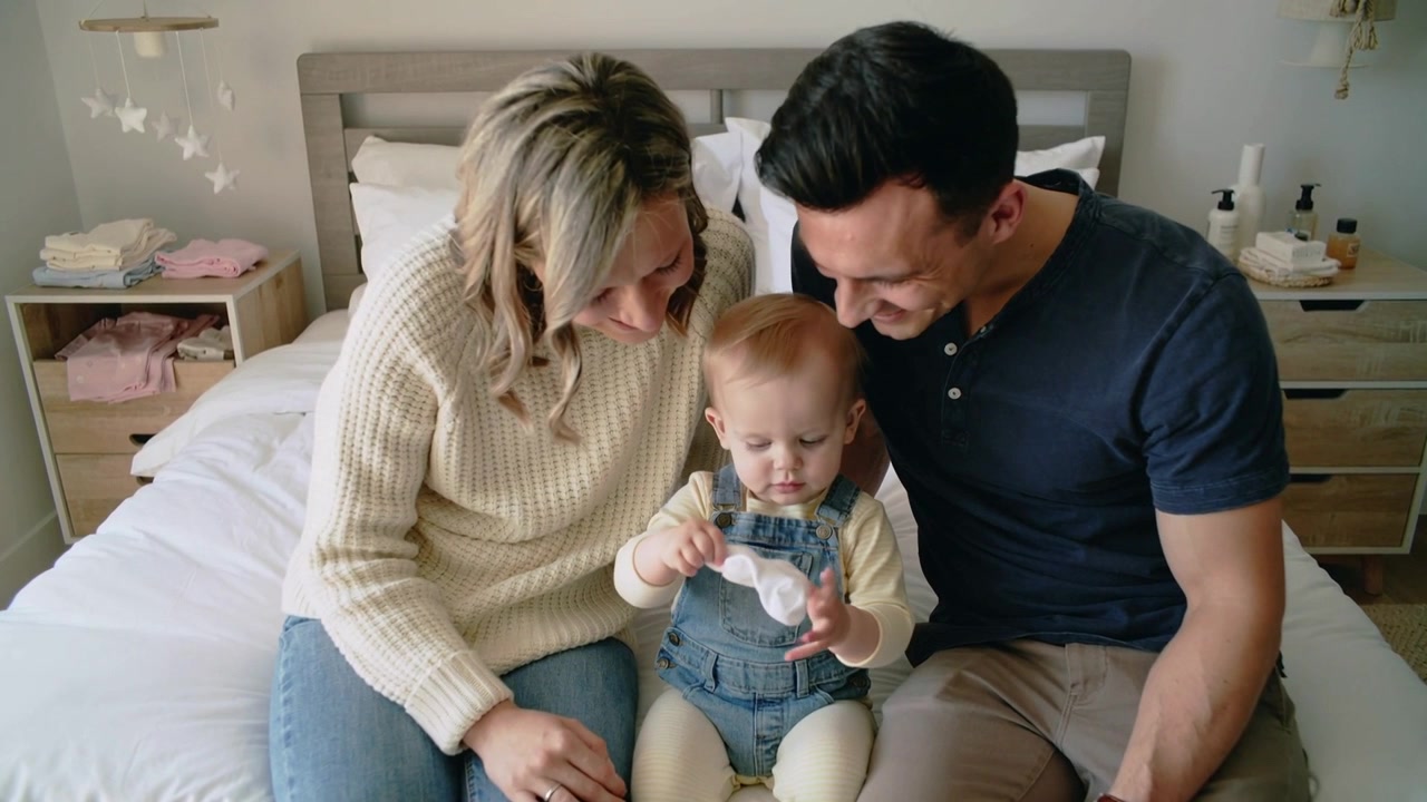 Mentre maneggia un piccolo panno bianco, un bambino piccolo in salopette lo esamina sul letto, curioso, con i genitori che sorridono.