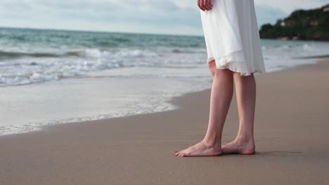 Close-up-of-young-woman-in-white-dress-bare-feet-on-the-beach,-waves-gently-touching-the-shore,-in-slow-motion