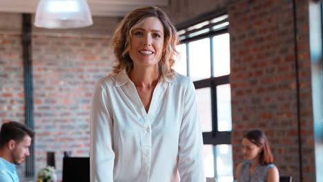 Portrait-Of-Smiling-Businesswoman-In-Office-With-Colleagues-Working-In-Background